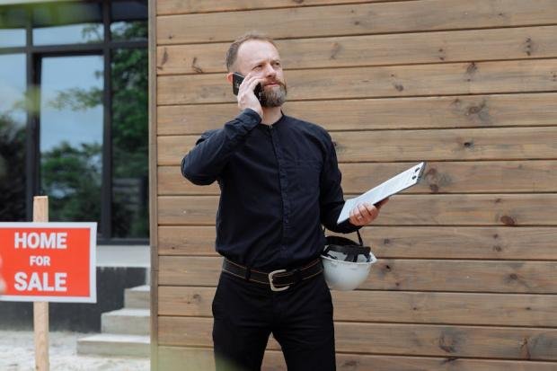Real estate agent making a phone call while holding documents near home for sale sign.