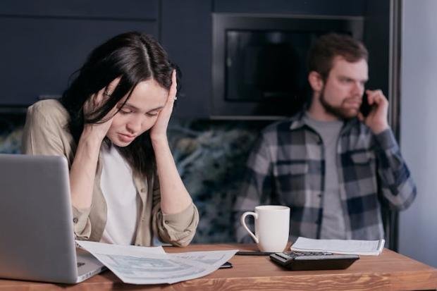 A stressed woman holds her head while working at a desk with documents and a laptop.